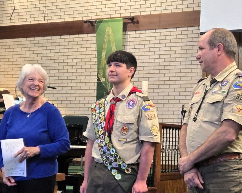 A boy scout is standing in a room with two men and a woman.