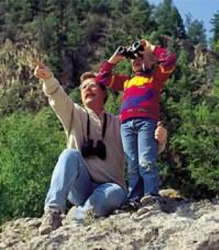Man and child birdwatching, one pointing, other using binoculars in a wooded area.