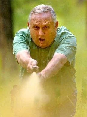 Man in green shirt fires a handgun, outdoors. Yellow smoke, surprised expression.