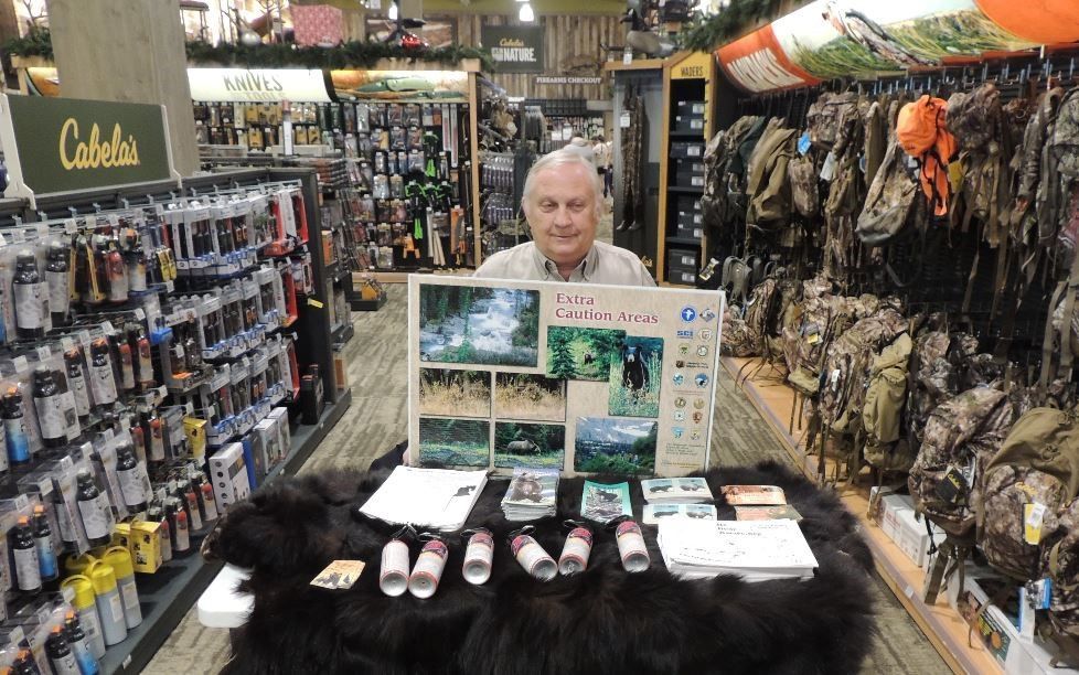 Man at Cabela's store display about hunting. Holds sign, surrounded by equipment & a bear skin rug.