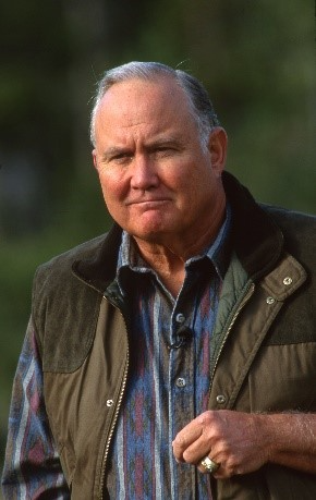 Man in vest and plaid shirt; outdoors, slightly smiling.