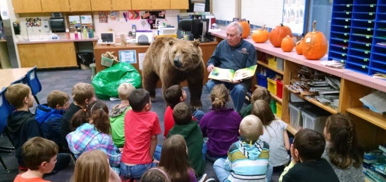 A man reading a book to children in a classroom with a large bear standing nearby.