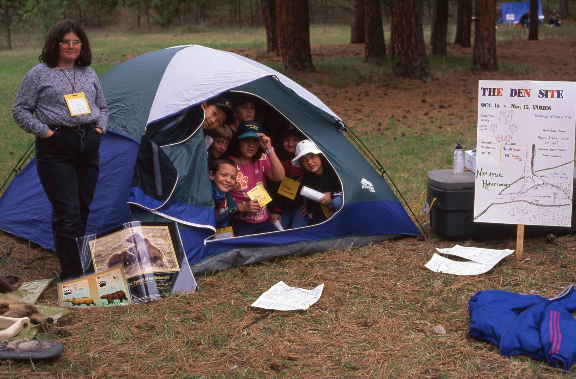 Woman and group of children in a tent, outside. Sign and maps nearby.