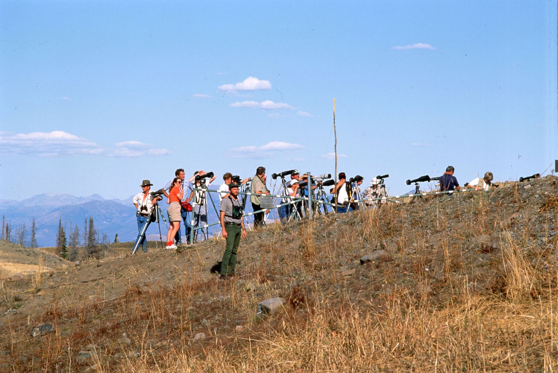 A group of people with binoculars and spotting scopes on a hilltop, observing a distant landscape under a blue sky.