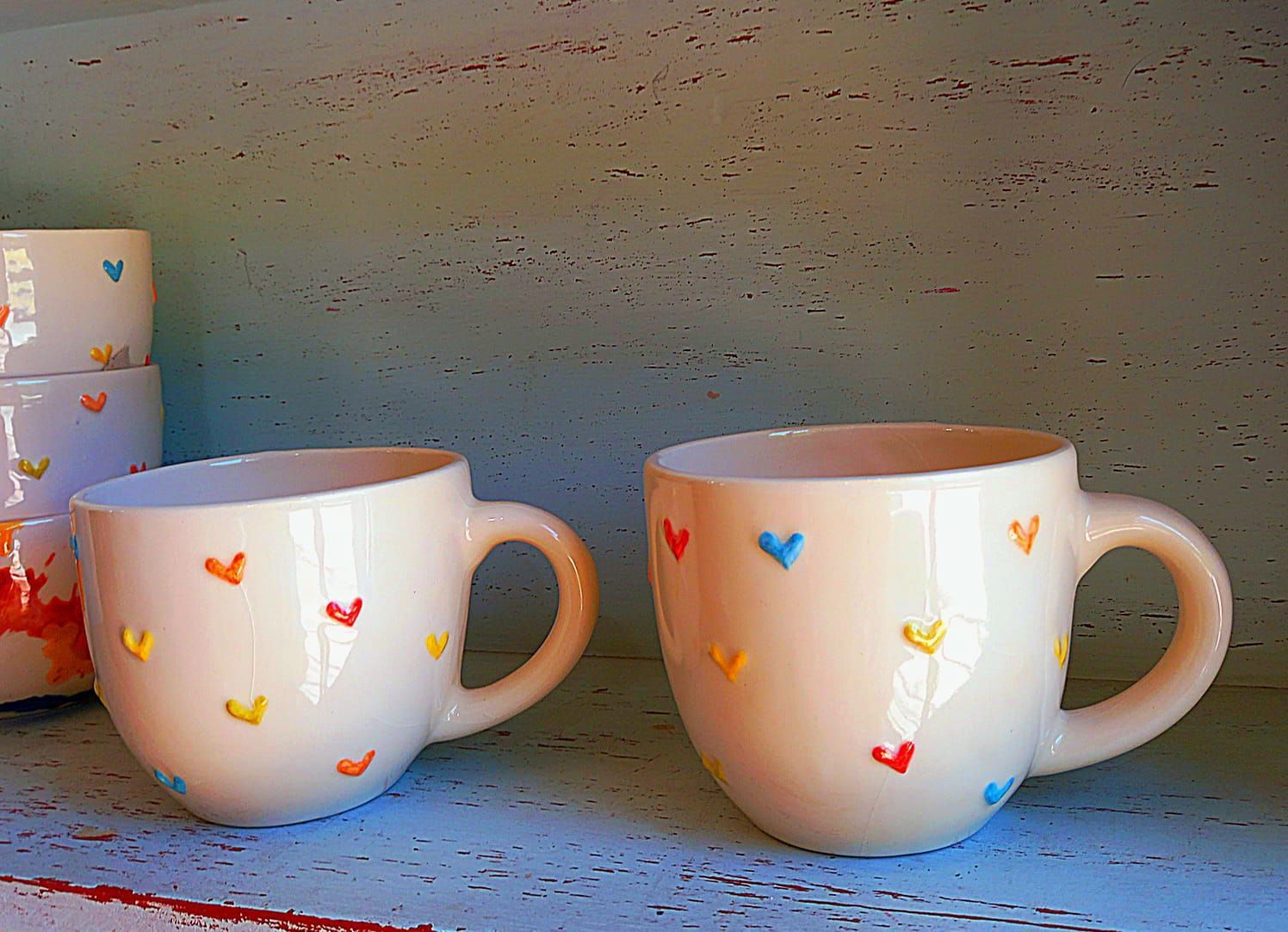 Dos tazas blancas con corazones sobre ellas se encuentran en un estante.