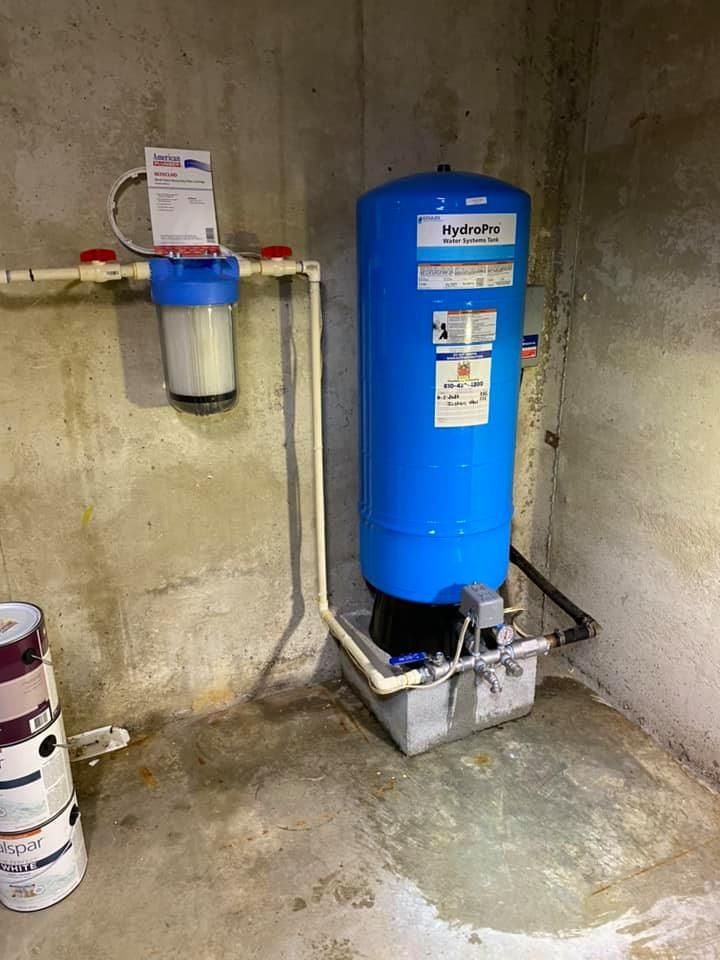 A blue water tank is sitting in a basement next to a bucket of paint.