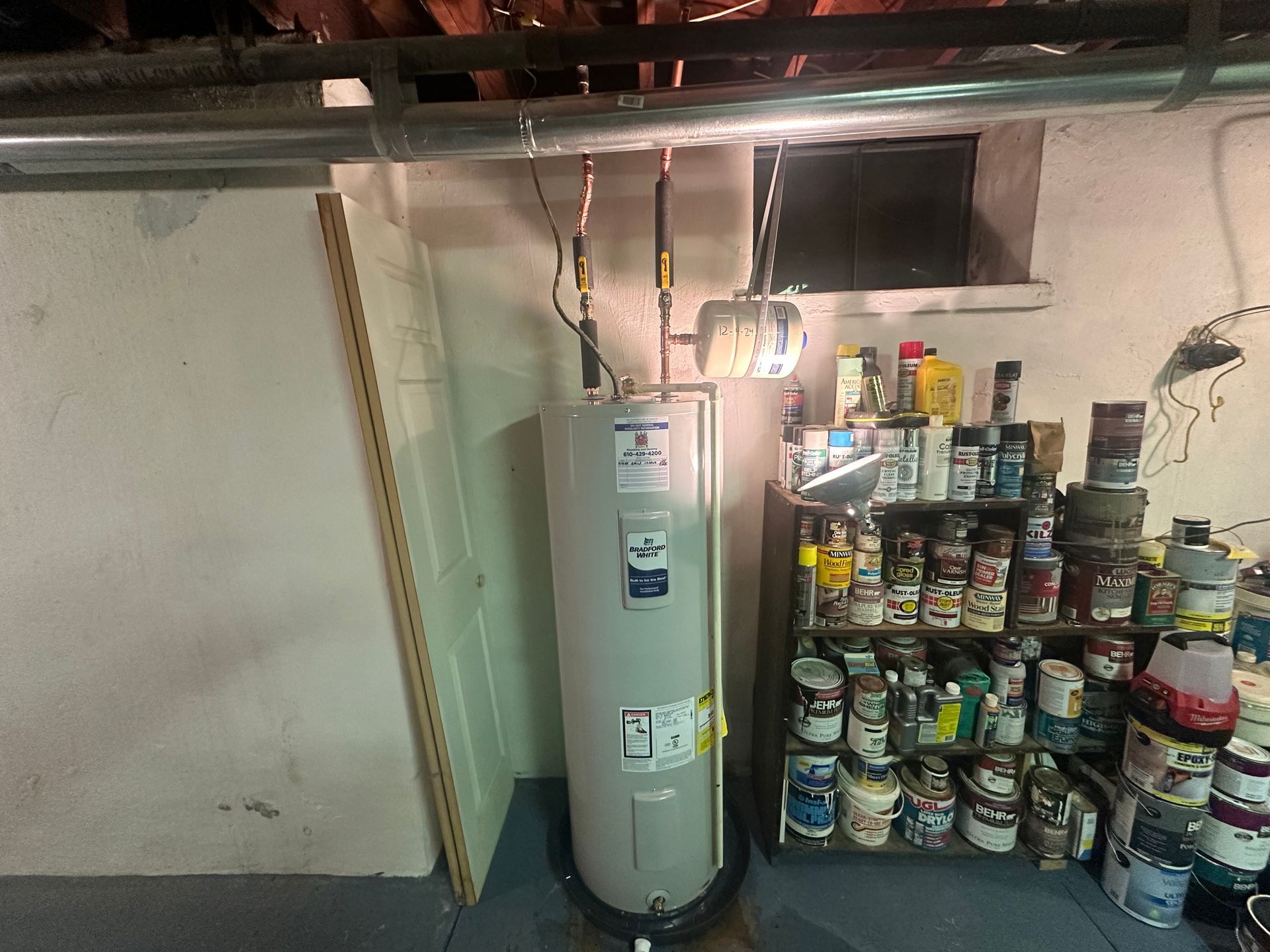 A water heater is sitting in a basement next to a shelf filled with paint.
