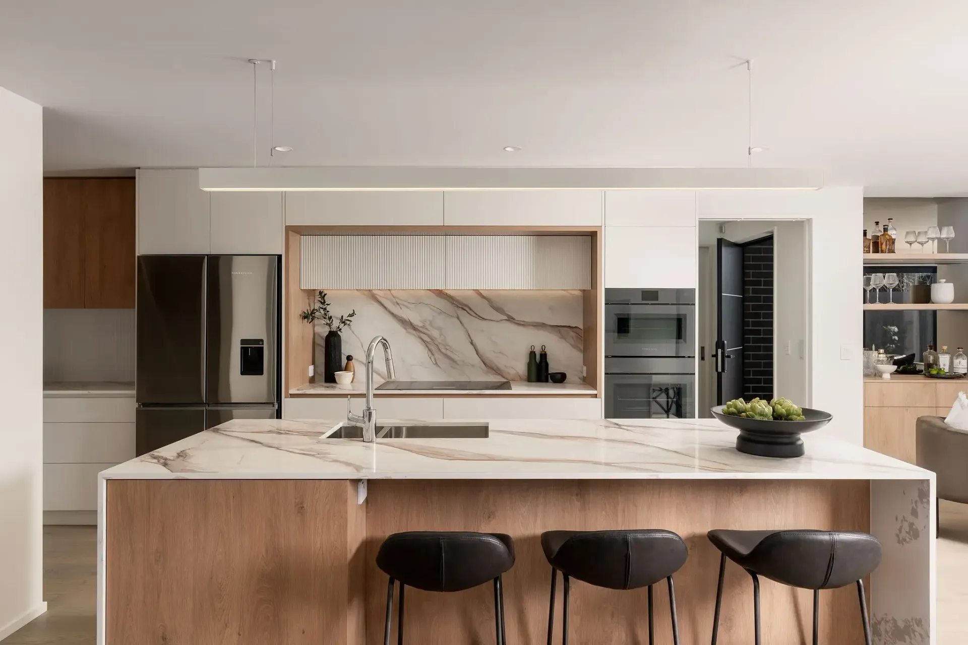 Modern kitchen with island, bar stools, light wood cabinetry, marble countertops, stainless steel appliances.