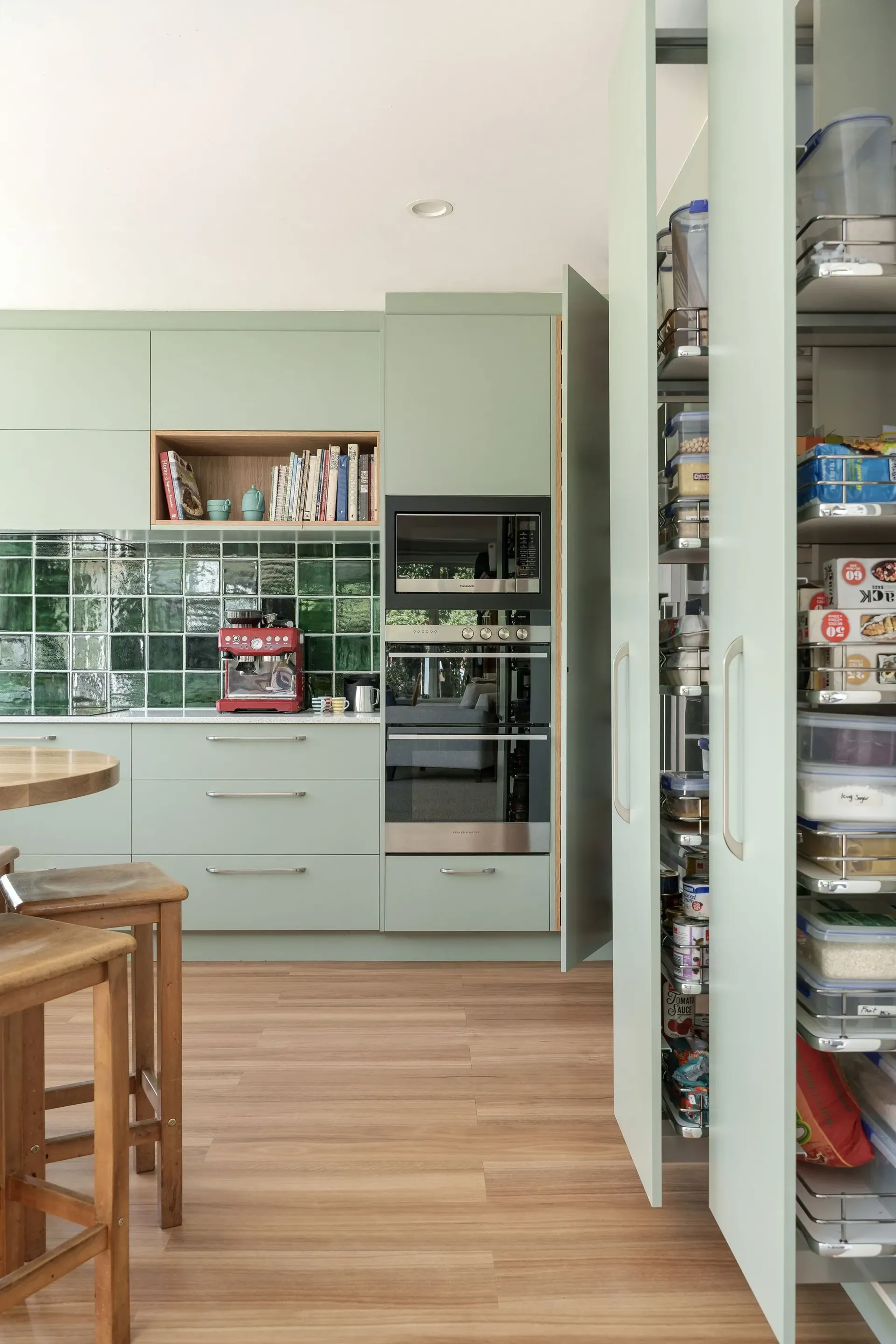 A kitchen with green cabinets , wooden floors , stools and a large pantry.