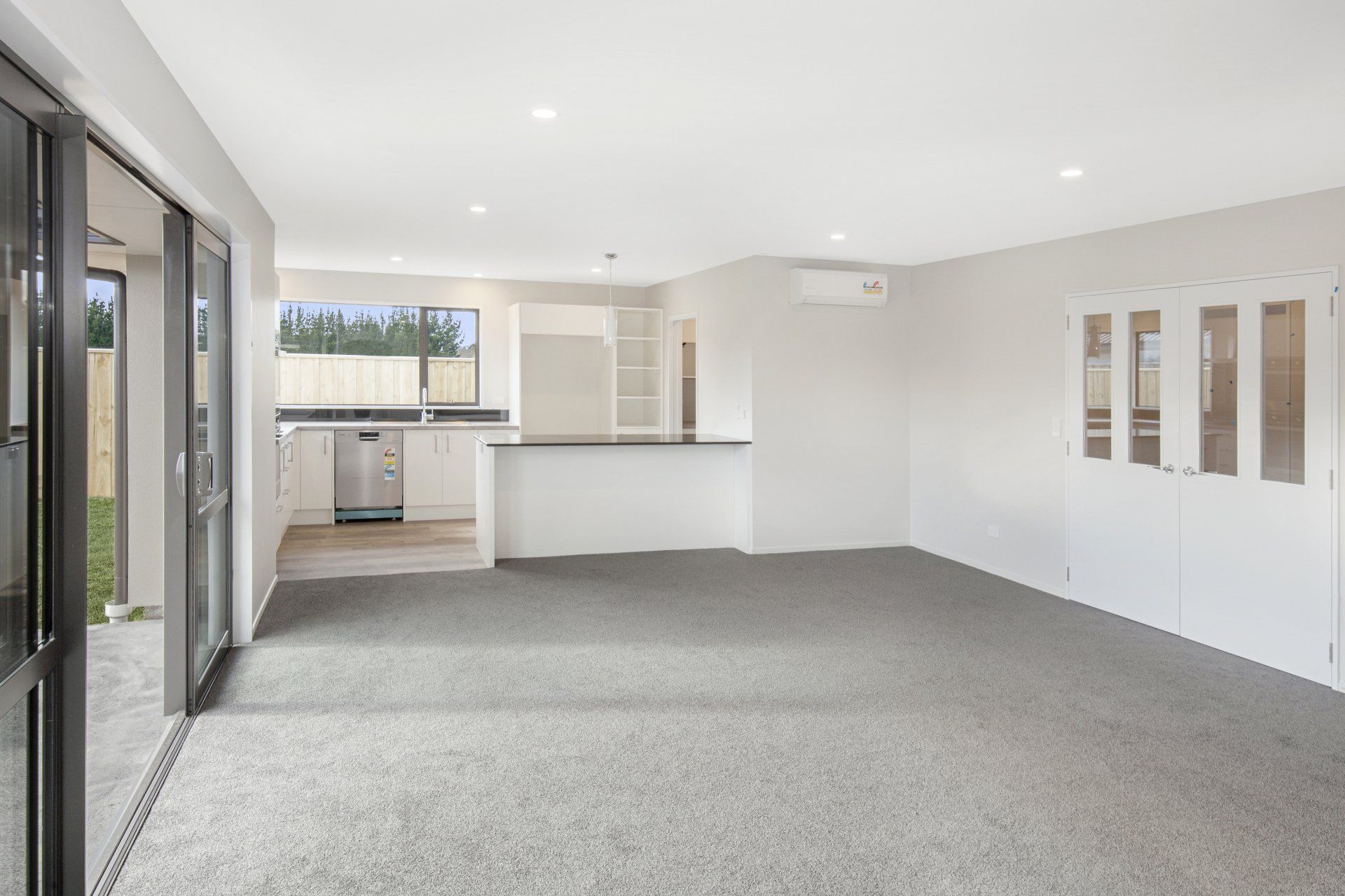 An empty living room with a carpeted floor and a kitchen in the background.