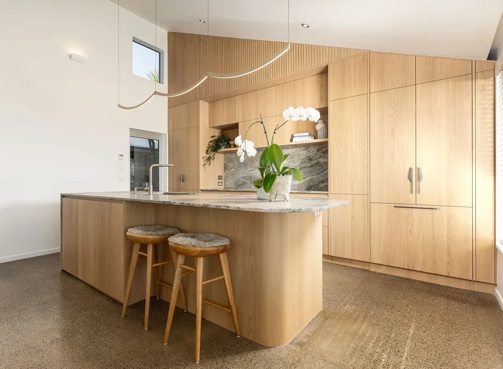 Modern kitchen with wood cabinetry, marble countertops, and bar stools; orchid plant.