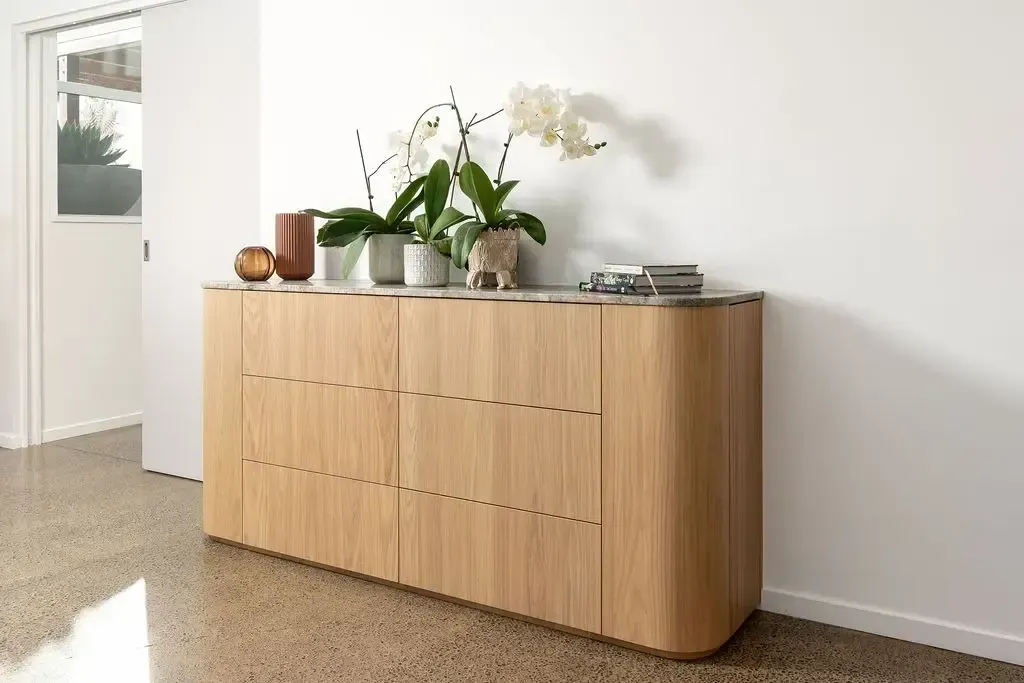 Wooden cabinet with curved edges against a white wall, topped with plants and decor.