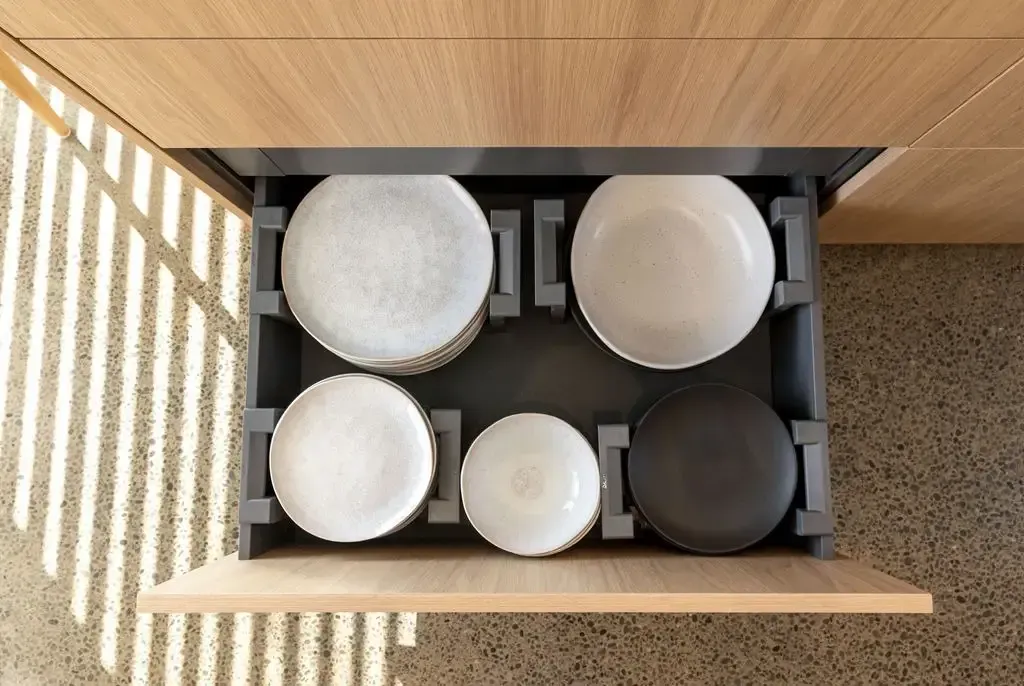 A drawer with gray dividers holds various white and dark plates, inside a light wood cabinet.
