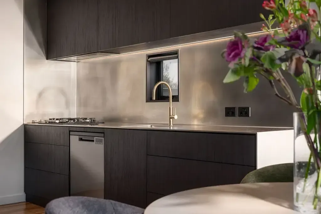 Modern kitchen with dark cabinets, stainless steel backsplash, gold faucet, and flowers.