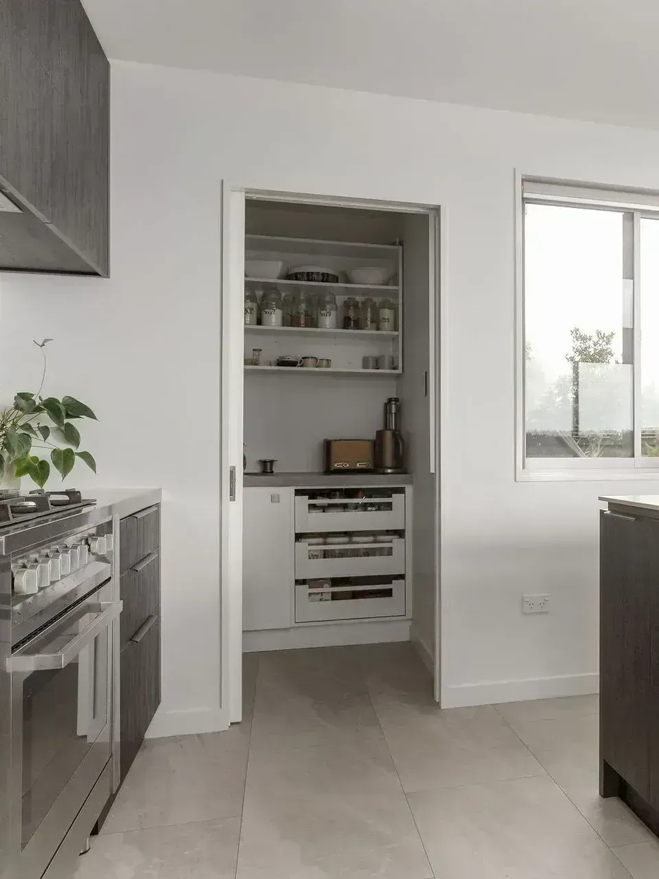 Kitchen with walk-in pantry. White walls, light-colored floor. Pantry has shelves and appliances. Window to the right.