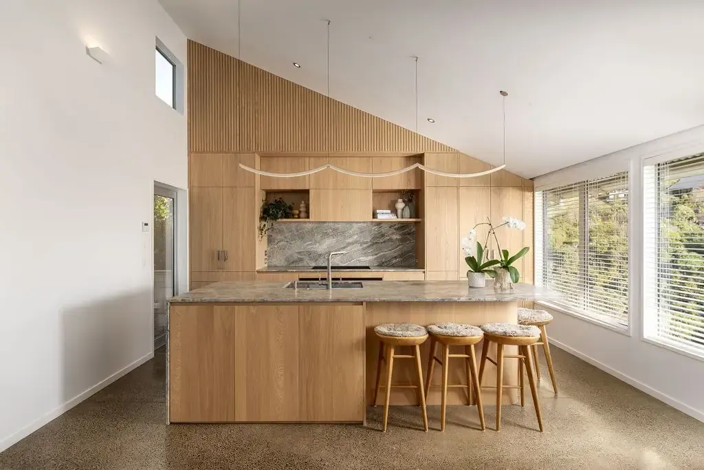 Modern kitchen with wooden cabinets, island, and stools, marble backsplash, and large windows.