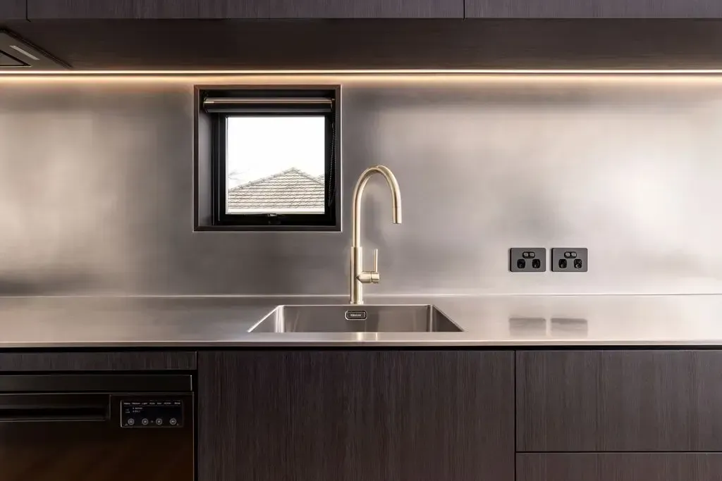 Modern kitchen: metallic backsplash, gold faucet, small window, dark cabinets, and a dishwasher.