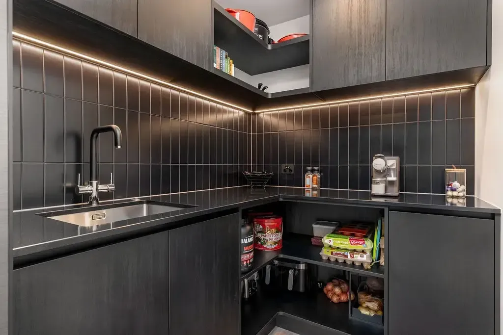 Dark modern pantry with black cabinets, shelves, and backsplash. Has sink and counter space, and some food visible.