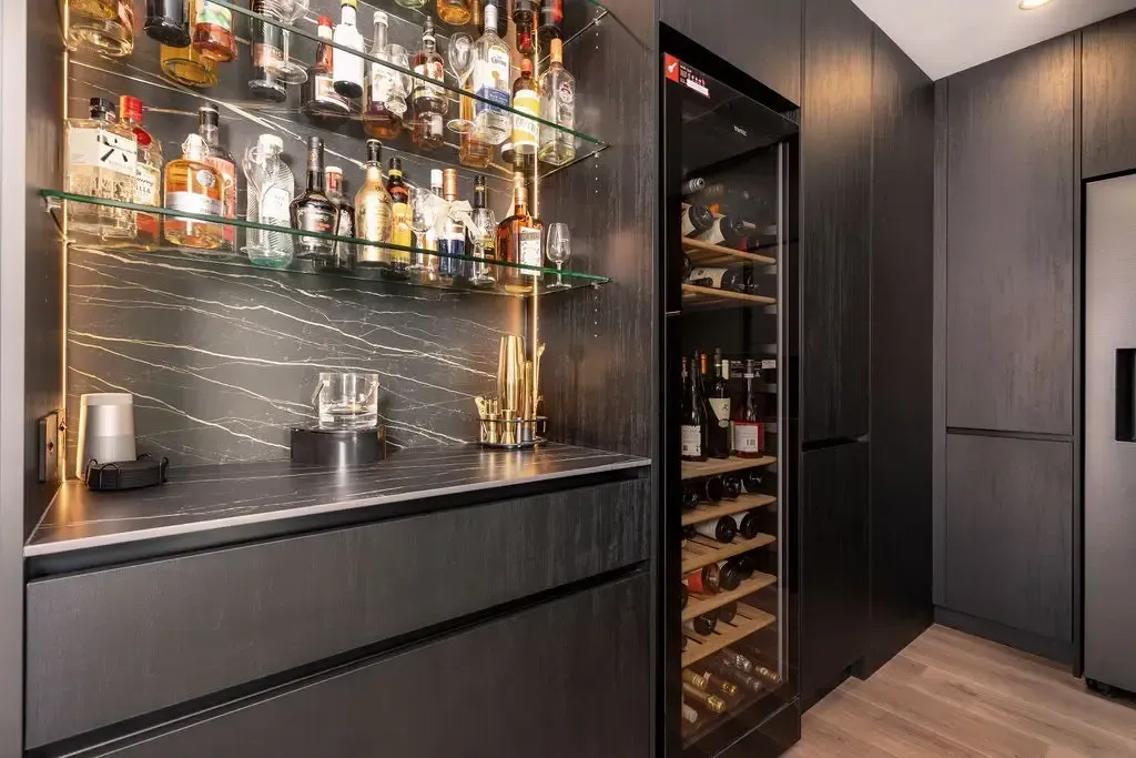 A modern home bar with liquor bottles on shelves, a wine fridge, and dark gray cabinetry.