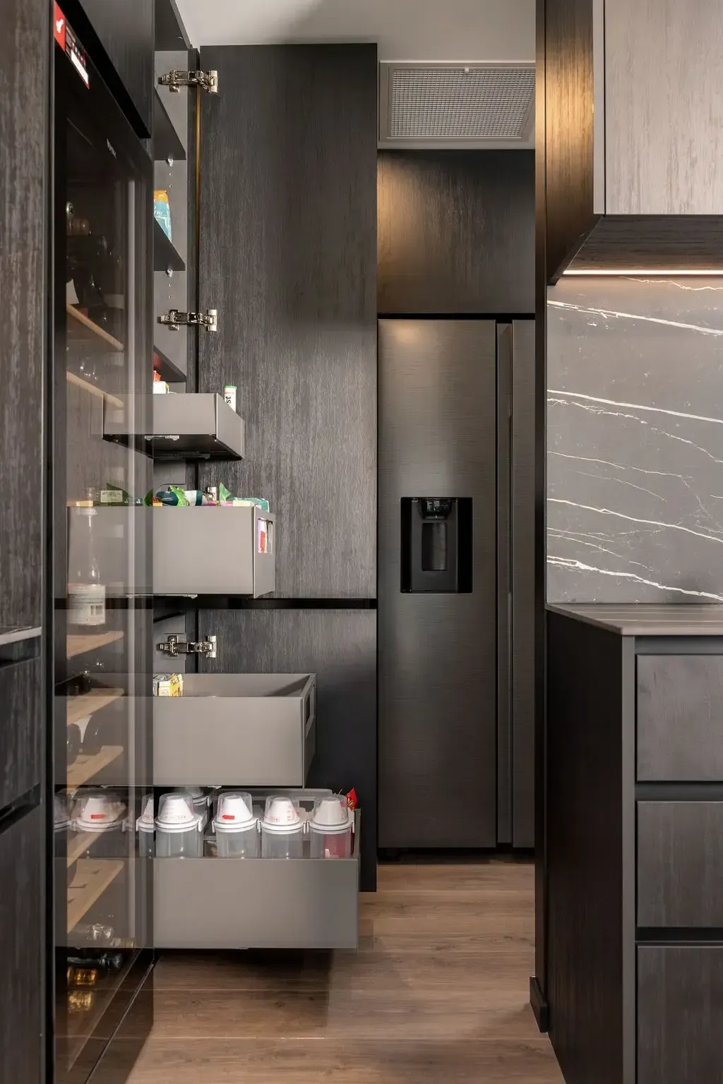 Dark kitchen interior: pantry with pull-out drawers, tall fridge, and wood cabinetry.