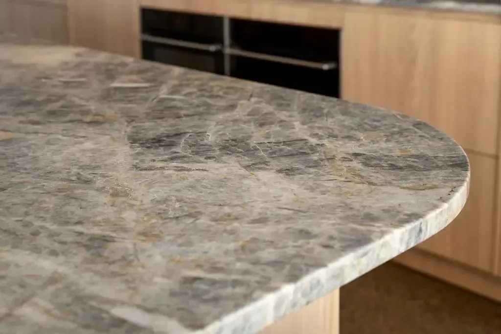Close-up of a marble countertop with rounded edge, light wood cabinets and oven in background.