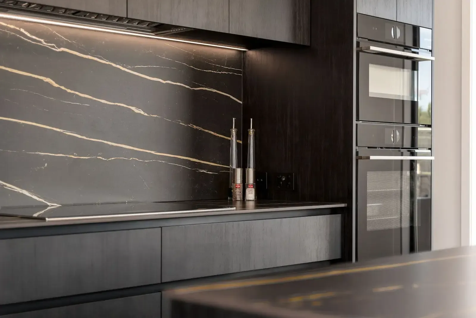Dark modern kitchen with black cabinets and countertop, gold veined backsplash, built-in ovens, and sleek design.