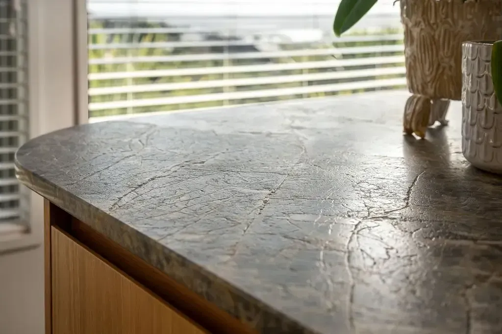 Closeup of a kitchen countertop with a textured gray surface. Wooden cabinet below, window with blinds in background.