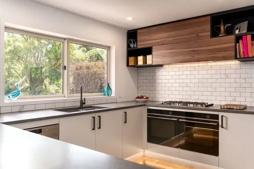 Modern kitchen with white cabinets, brick backsplash, wood accents, and a large window.