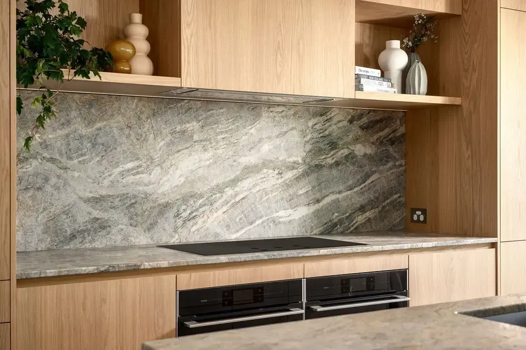 A modern kitchen with light wood cabinets, gray stone backsplash, and built-in ovens.