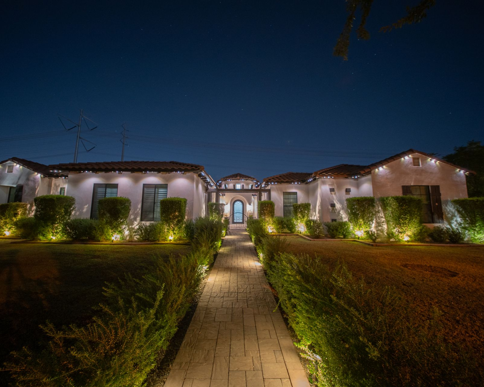 Night view of a house with illuminated pathway, bushes, and decorative lights.