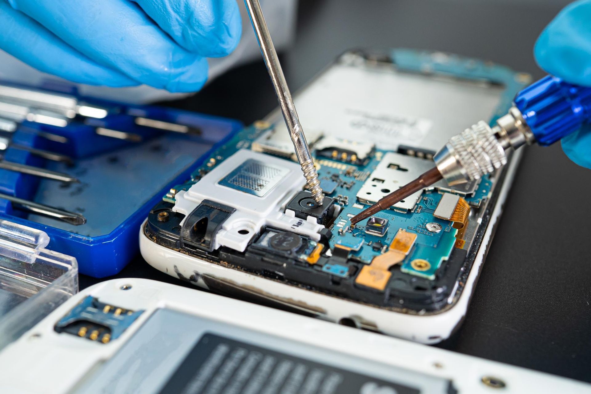 Technician repairing a smartphone motherboard with tools and blue gloves on a workbench