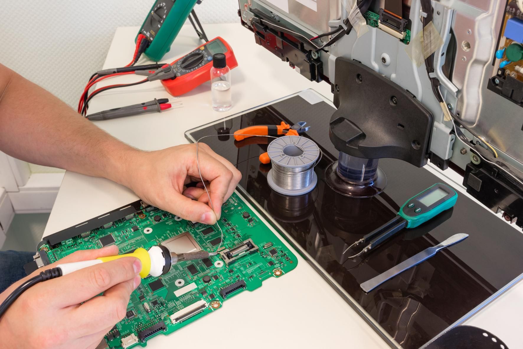 Hands repairing a green circuit board on a workbench with tools and laptop nearby