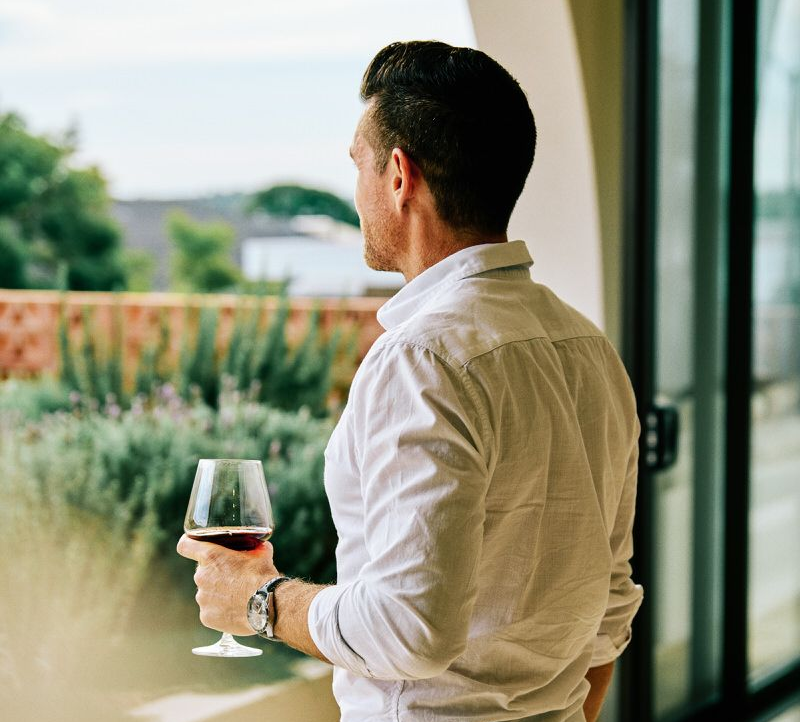 Man in a white shirt holding a wine glass, looking out a window at a garden view