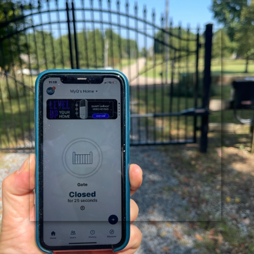 Open metal gate on a paved path, leading to a walkway with trees and buildings under a blue sky.