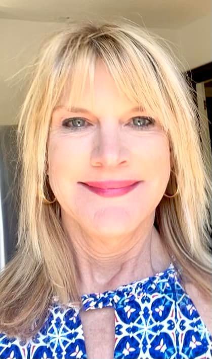 Blonde woman smiling, wearing a blue patterned top and gold hoop earrings.