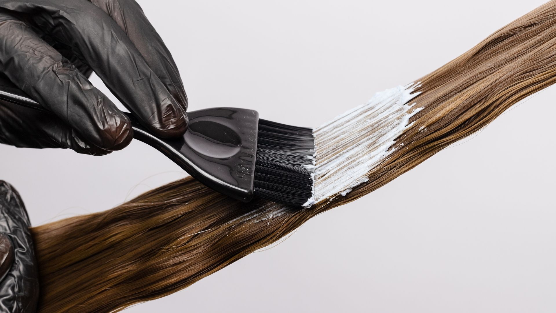 Person with black gloves applying hair dye with a brush to a strand of brown hair.