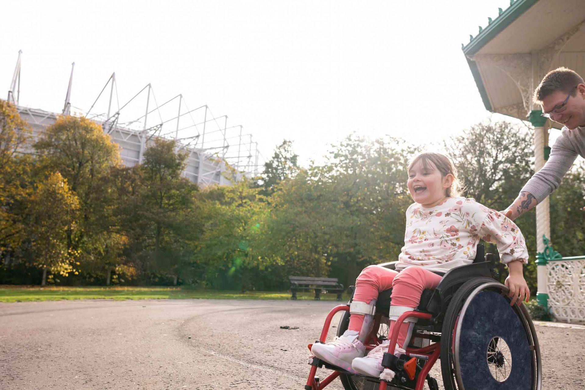 A smiling child in a wheelchair is pushed by a person outdoors in a park.