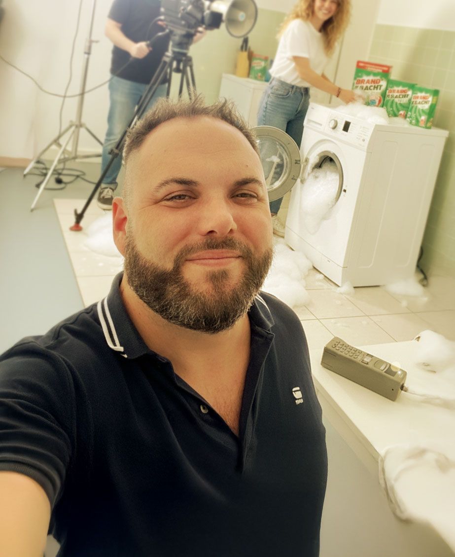 Man die een selfie maakt, wasmachine vol schuim, filmploeg op de achtergrond met producten.