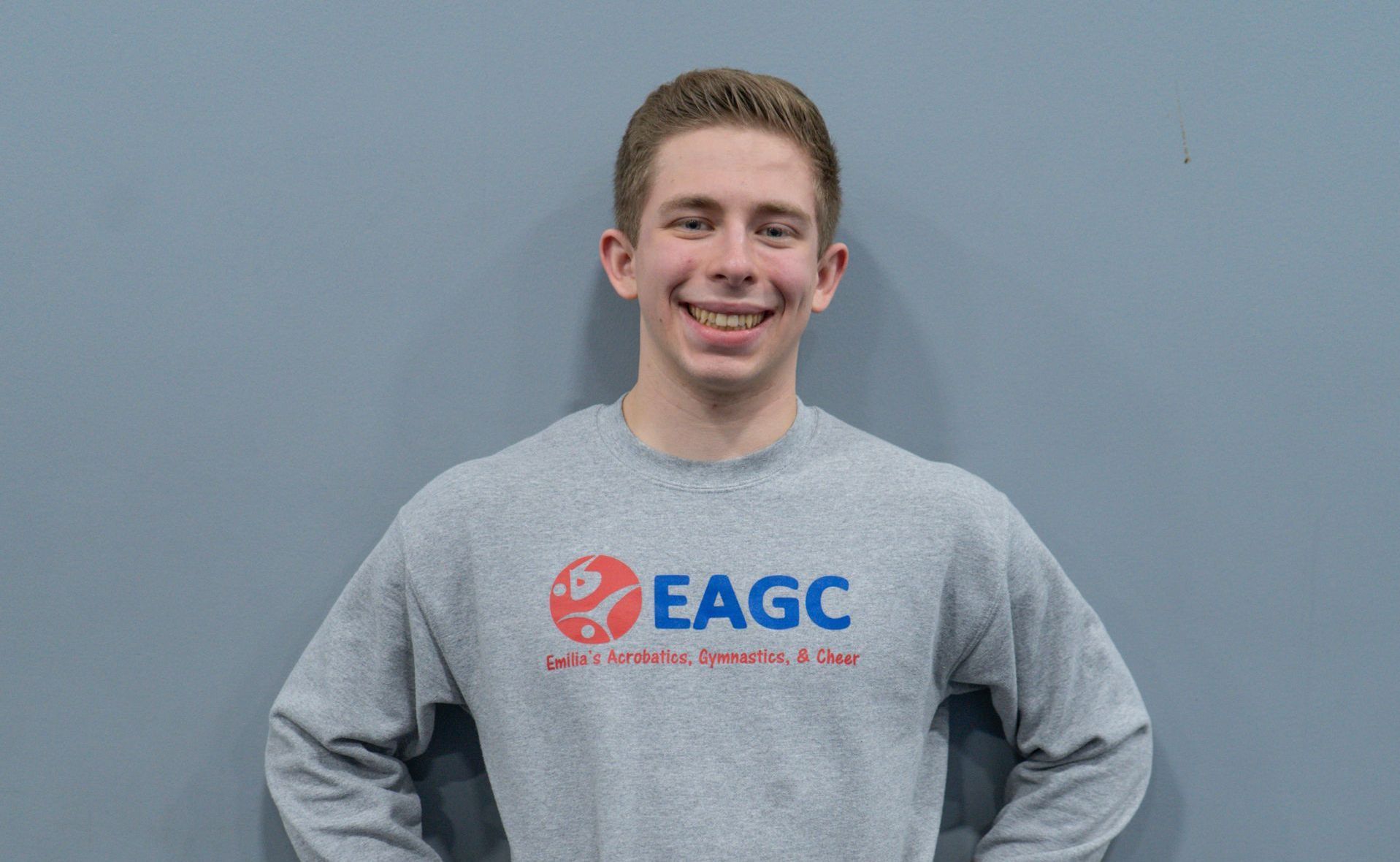 A young man wearing a gray eagc sweatshirt is standing in front of a blue wall.