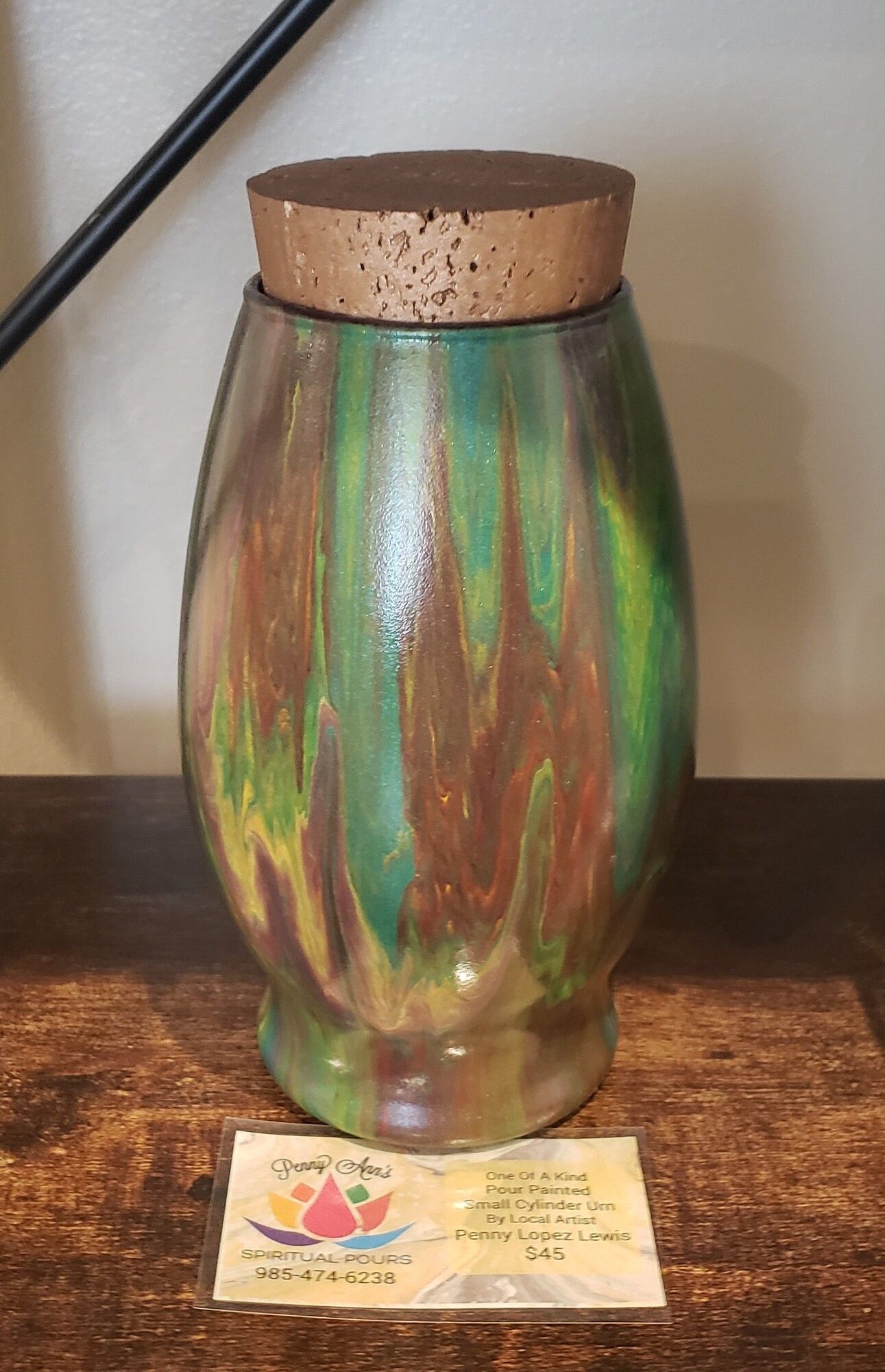 Wooden jar with swirled green, brown, and yellow paint and a cork lid, sitting on a table with a business card.