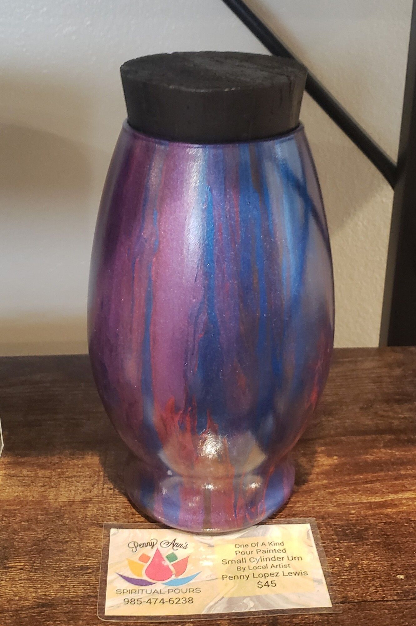 Purple and blue painted urn with black lid, on a wooden surface, with a business card in front.