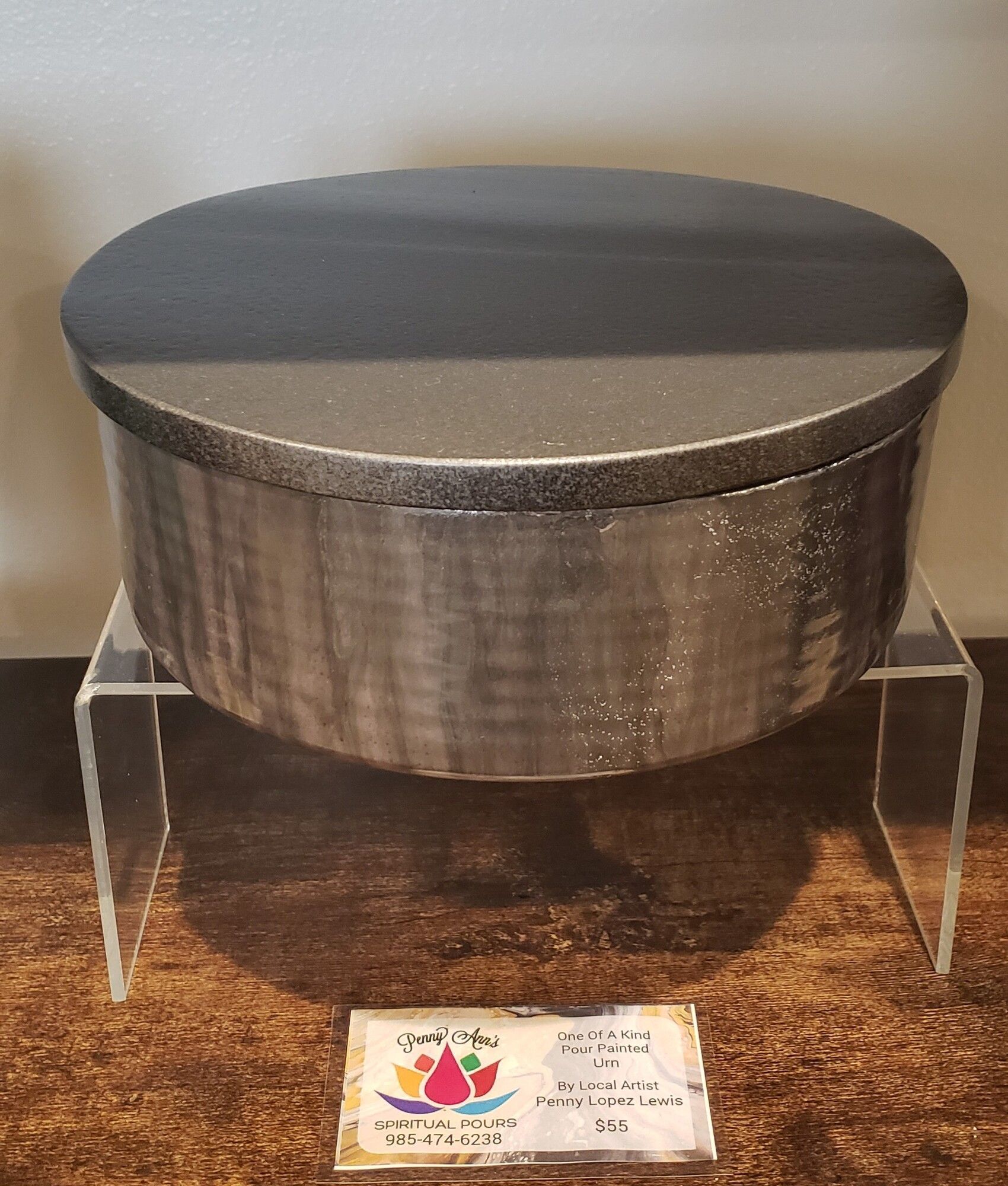 Silver urn with a black lid, on a clear acrylic stand, against a white wall.