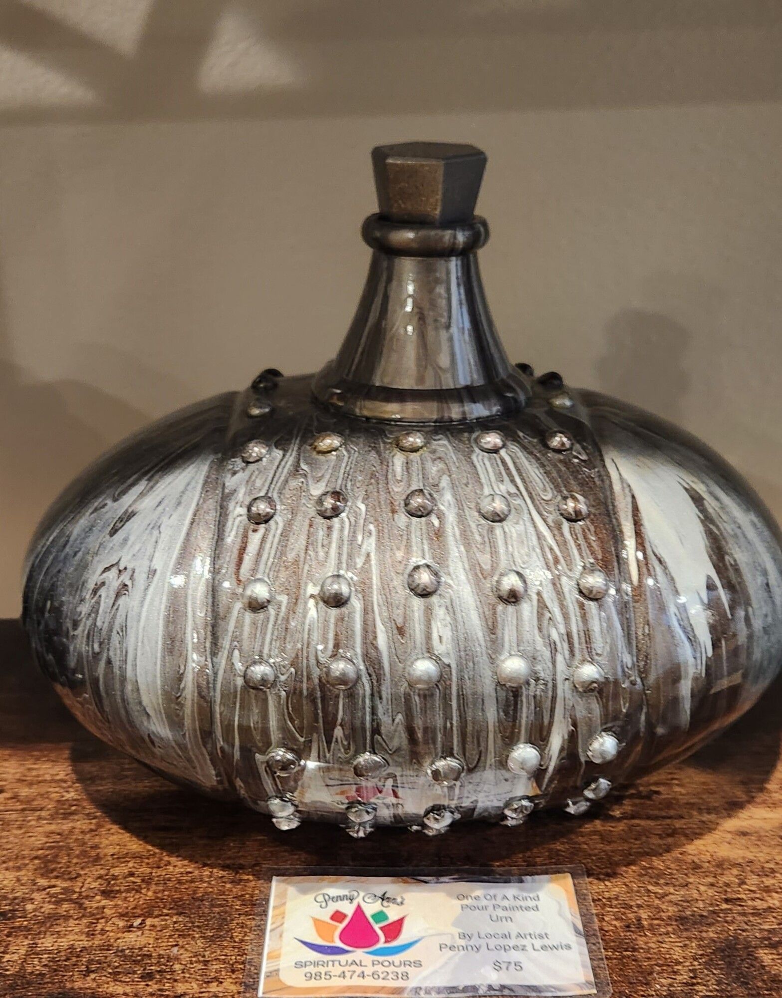 A bulbous, metallic-toned vase with raised dots, on a wooden surface. The vase has a dark neck and a label at the base.
