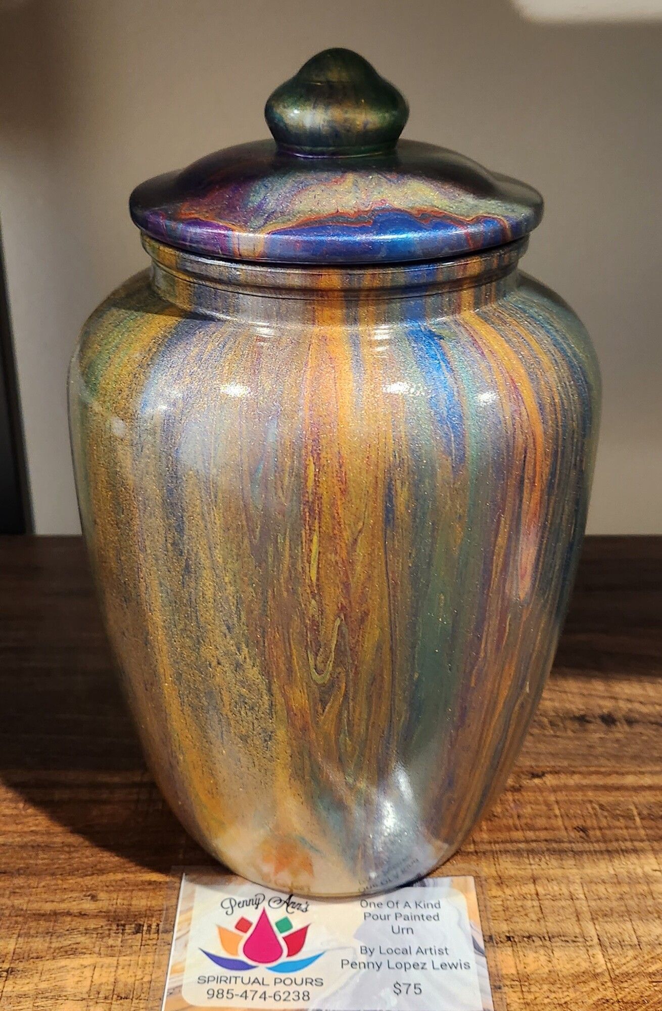 Metallic painted urn with lid, in shades of gold, blue, and purple, resting on a wooden surface.
