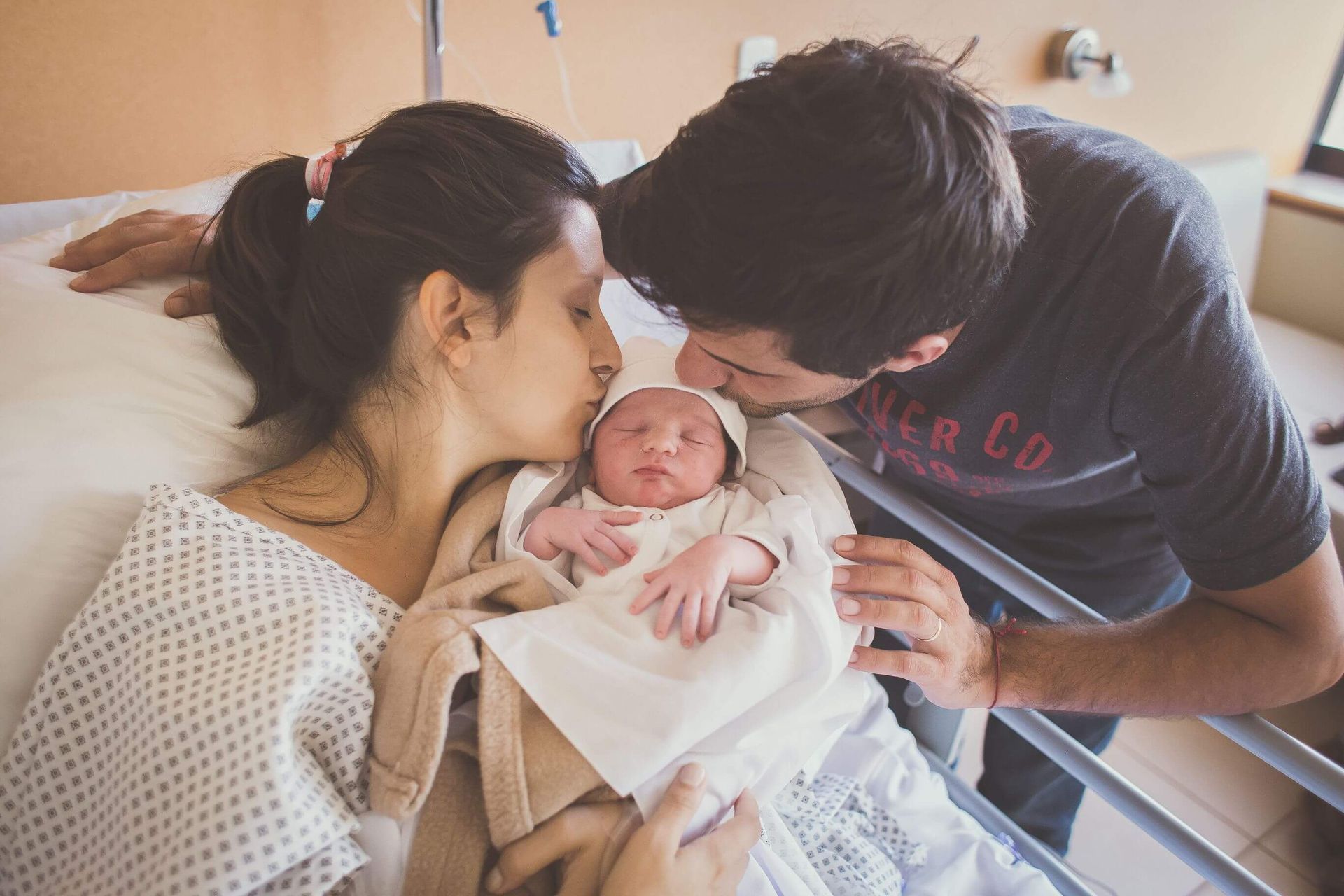 Los padres besan a un bebé recién nacido en una cama de hospital; el bebé está envuelto en una manta