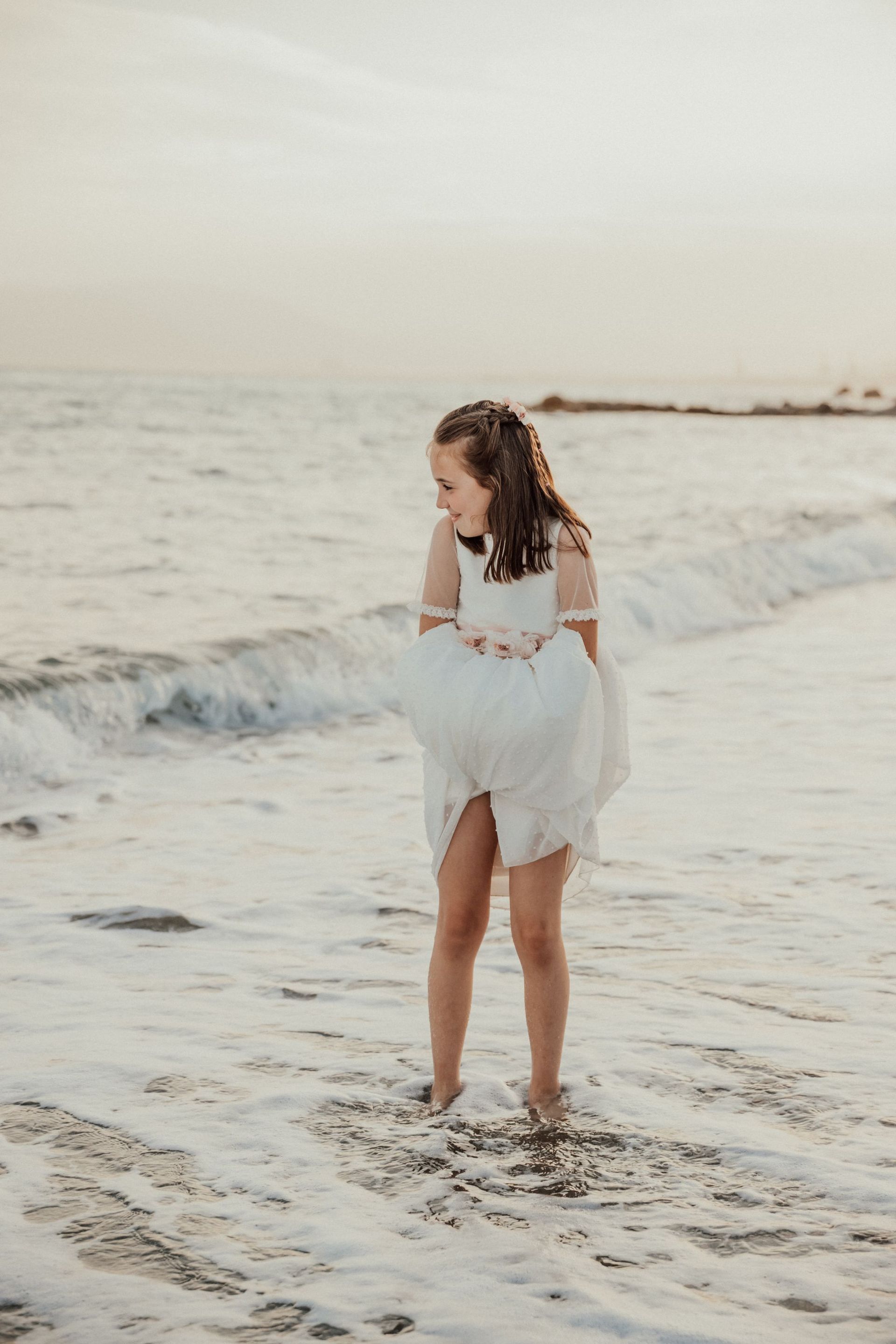 Una niña con un vestido blanco está parada en la playa mirando el océano.