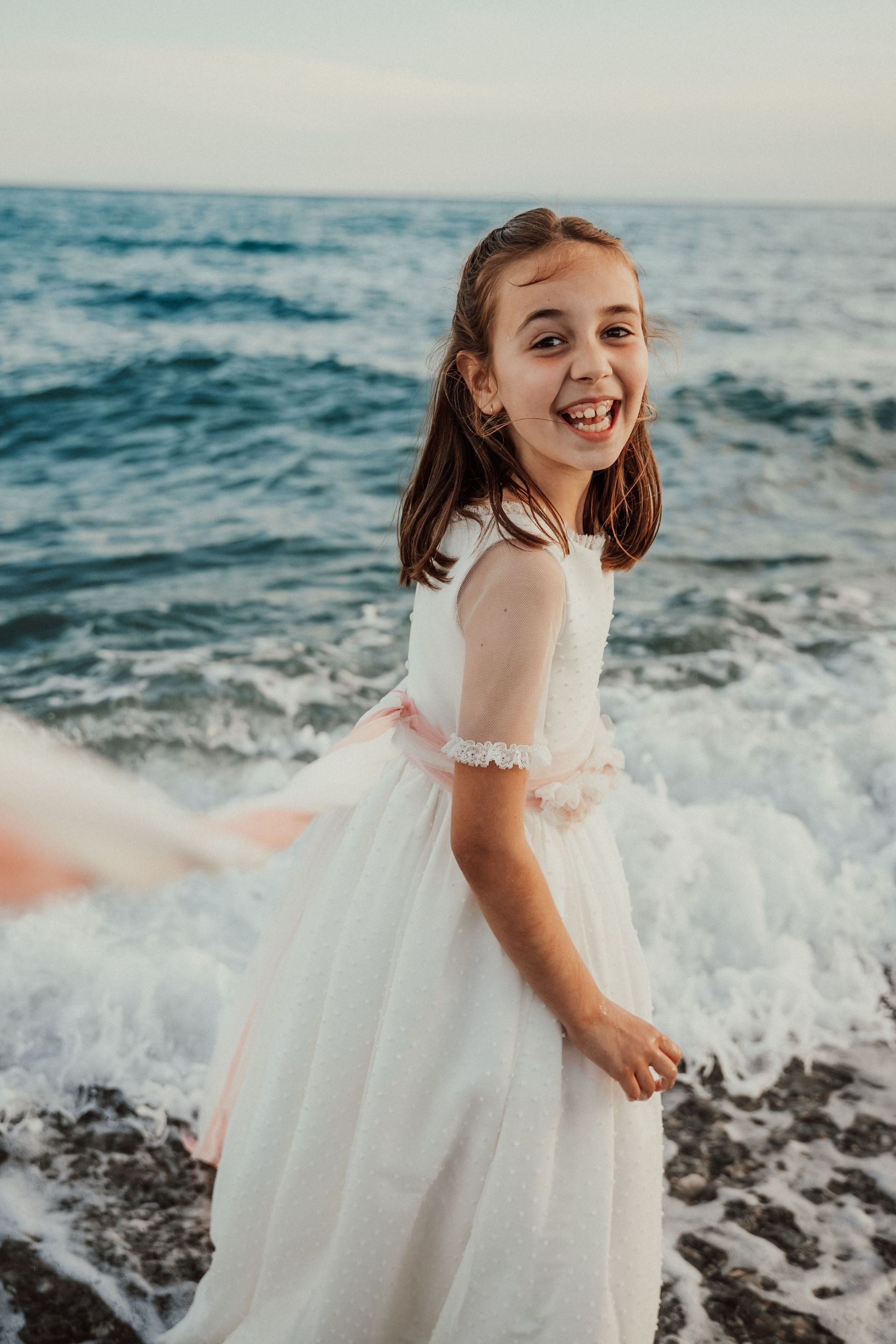 Una niña con un vestido blanco está parada en la playa cerca del océano.