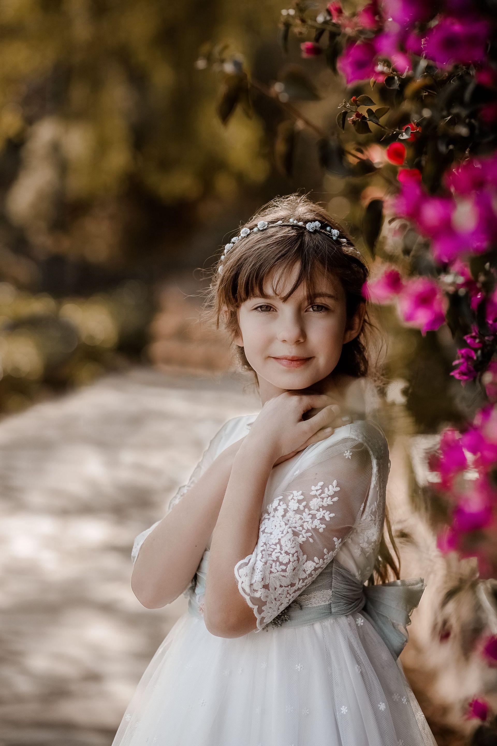 Una niña con un vestido blanco está parada junto a un arbusto de flores moradas.
