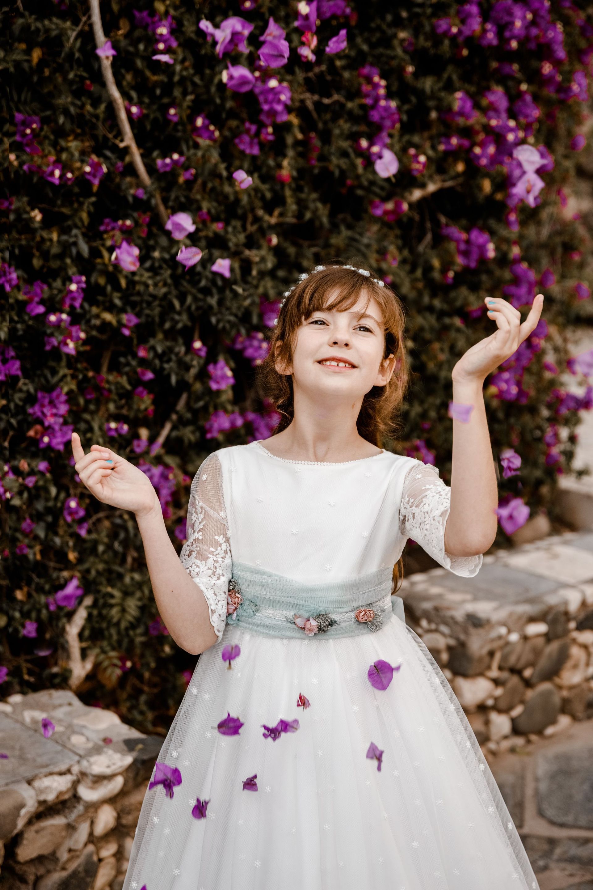 Una niña con un vestido blanco está parada frente a un arbusto de flores moradas.