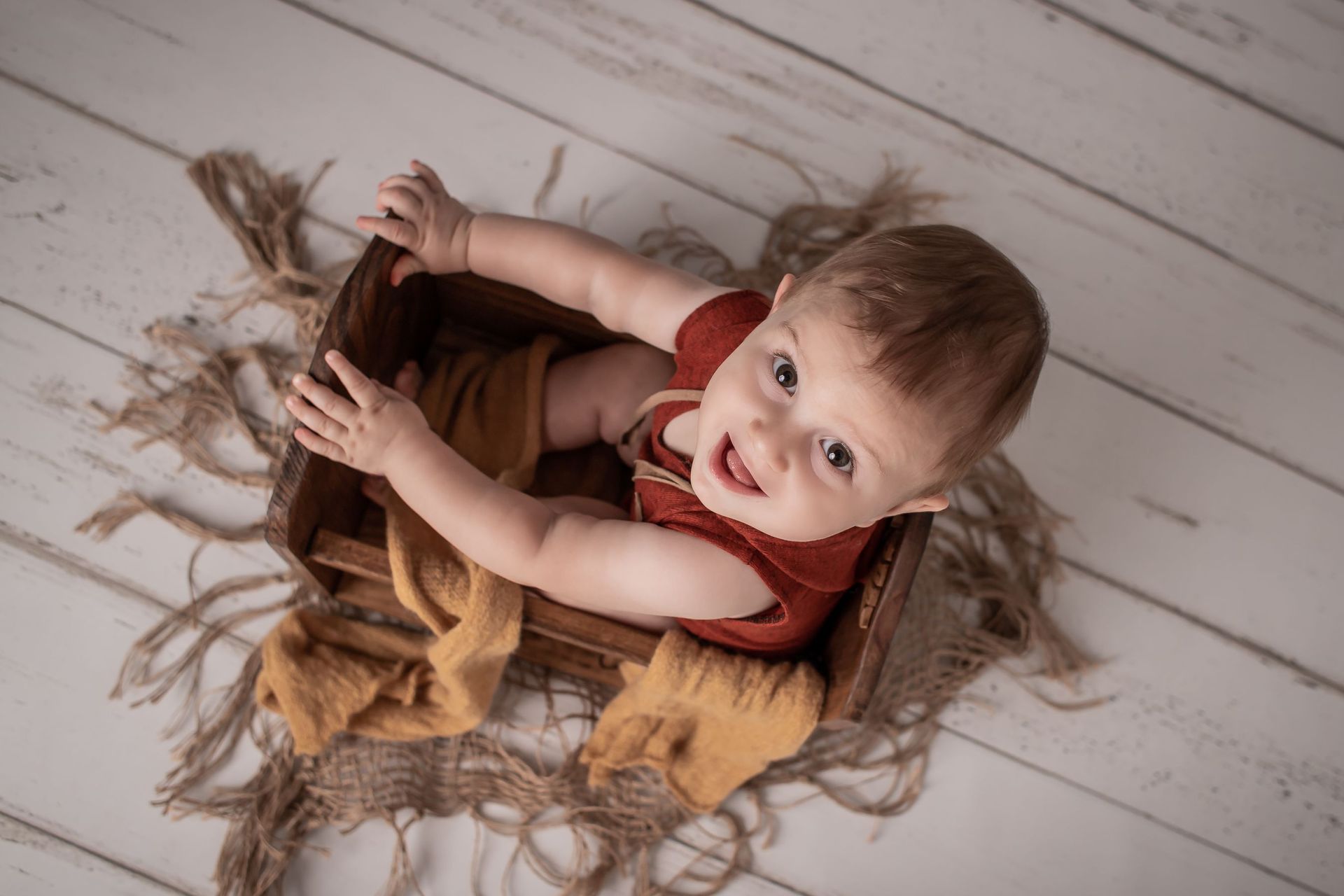 Bebé en una caja de madera, sonriente, vestido de rojo, rodeado de cuerdas y tela amarilla, sobre un piso de madera.